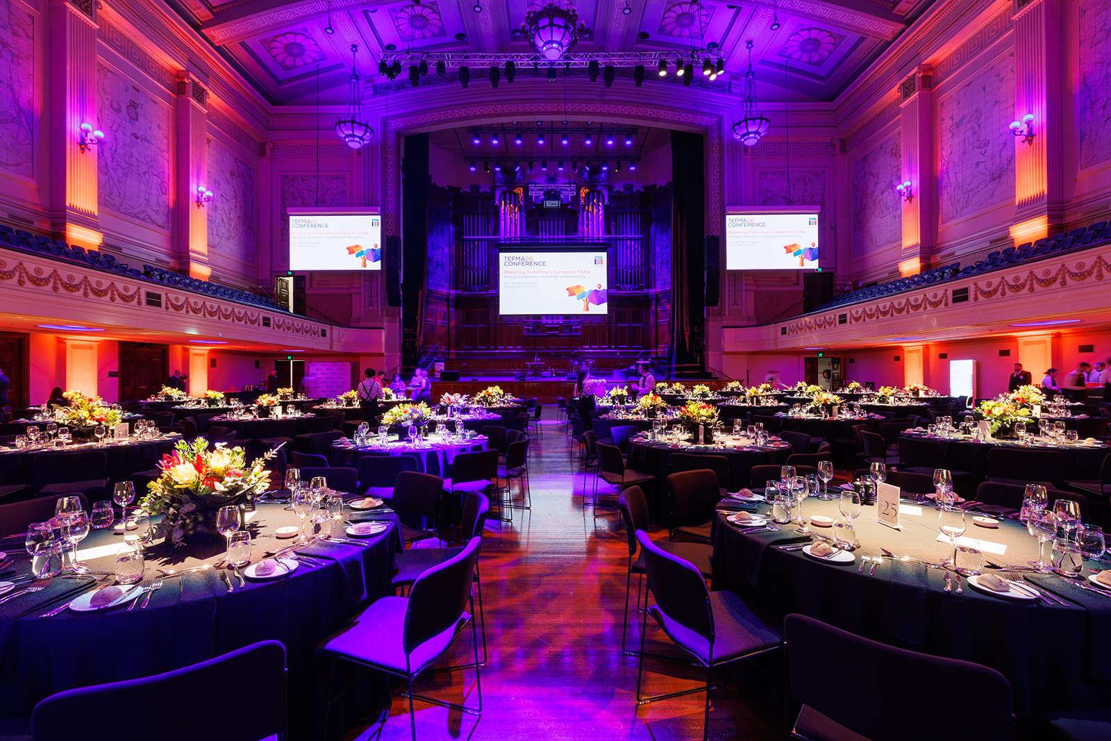 Delegates seated at round tables in a grand hall lit in violet for a conference gala dinner